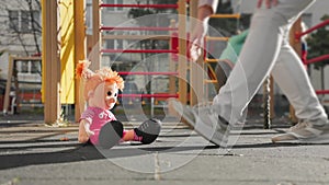 A toy doll sits on the ground on the playground. A woman passes by and takes a toy with her. Low angle view. Close-up