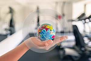 Toy globe in the female hand in fitness gym opposite the sport equipment and  jogging simulators. Healthy lifestyle. World health