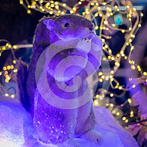 A toy gopher with raised paws against the background of an artificial forest and snow. Christmas and New Year concept