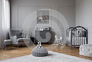 Toy on grey pouf in the middle of trendy baby bedroom with grey wooden cradle, chest of drawers and comfortable armchair, real