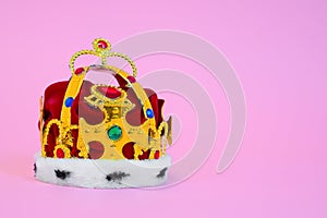 Toy plastic royal crown decorated with fake gems, fur and red cloth on pink background, studio shooting, front view