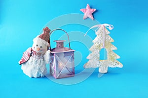 Toy snowman in a Santa hat, lantern with a lit candle on a blue bright background, Merry Christmas concep