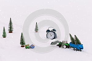 Toy tractor with a trailer carries Christmas trees during snowfall, rides through the snow in the middle of the forest.