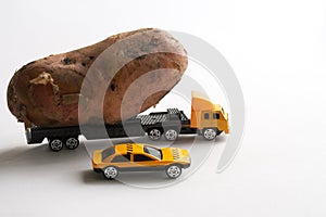 Toy truck, accompanied by a security car, carries a large potato. White background. The concept of delivery of oversized items and