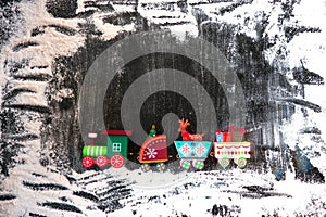A toy wooden train carries gifts and Christmas paraphernalia on a dark, snow-covered background. Image for a decorative Christmas
