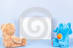 Toys with empty white frame on light blue background. Toy teddy bear, plush stuffed elephant and white wooden picture