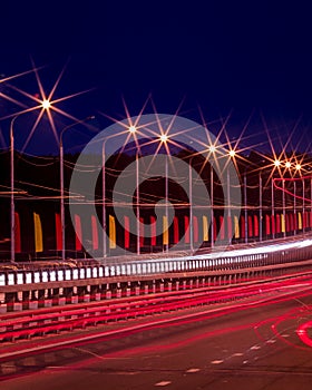 Traces of headlights from cars moving at night on the bridge, illuminated by lanterns. Abstract cityscape with highway at dusk