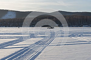 Traces from the wheels of cars lead along the snowy plain of the lake to the fishermen against the background of the coast.
