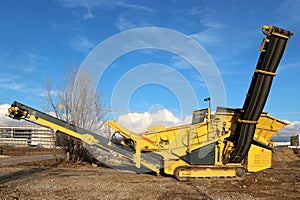 A tracked stone crusher machine under a blue sky with clouds