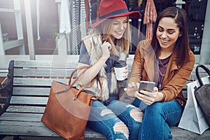 Tracking the sales online. two young girlfriends sending text messages while sitting on a bench during a shopping spree.
