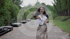 Tracking shot of confident elegant slim Asian woman walking on alley in summer park turning looking at camera smiling