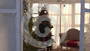 Tracking shot of decorated Christmas tree in room by frozen window on sunny day. Xmas tree decorated with lights and