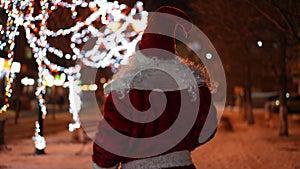 Tracking shot of positive relaxed Santa dancing walking on snowy night alley with Christmas light waving away. Back view