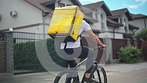 Tracking shot of young Caucasian man with yellow delivery bag riding bicycle on suburban street. Live camera follows