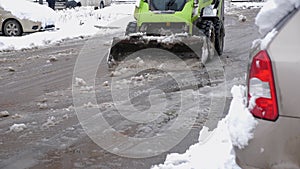 A tractor with a bucket cleans the road in the parking lot from slush and snow.