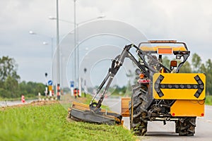 Tractor with a mechanical mower mowing grass on the side of the asphalt road