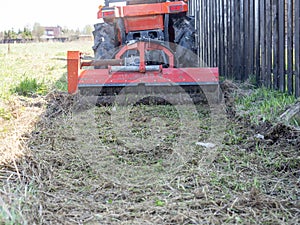 A tractor with a mower attached mulches dry grass along the fence. Land plot processing