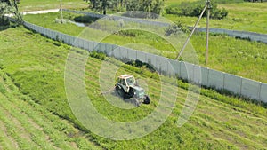 Tractor mows green grass in a meadow near a fence: haymaking