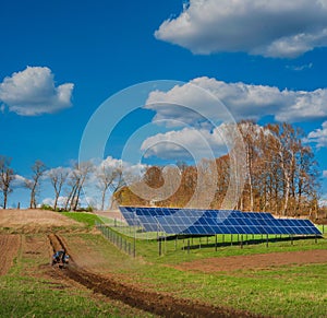 Tractor plows on a spring field and near the solar panel systemand clouds in the sky