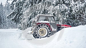 Tractor removing snow from large snowbanks next to forest road, after blizzard in winter