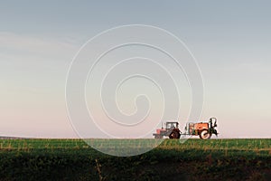 Tractor spraying pesticides on field with sprayer at sunset