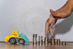 Tractor toy downloading a coins stack, hand as finger running on heap of coins