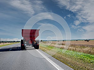 Tractor with trailer on a road by a big agriculture field. Warm sunny day with blue cloudy sky. Food supply chain. Grain product