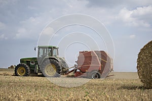 A tractor uses a trailed bale machine to collect straw in the field and make round large bales.