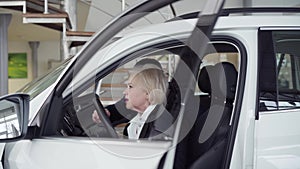 Trader helping senior female client to examine car interiors in dealership. Senior blond woman and adult man sitting in