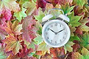 Traditional alarm clock on a background of green, yellow, orange, and red maple leaves, fall time change concept