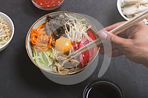 Traditional Asian Bibimbap dish with rice and vegetables on dark background. Man takes the dish using chopsticks