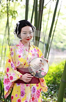 Traditional Asian Japanese beautiful Geisha woman wears kimono hold a fan on hand under a tree in a summer graden