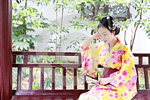 Traditional Asian Japanese beautiful Geisha woman wears kimono hold a fan sit on a bench in a summer nature garden