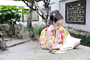 Traditional Asian Japanese beautiful woman wears kimono with fan on hand reading book in outdoor spring garden