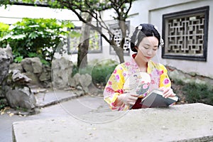 Traditional Asian Japanese beautiful woman wears kimono with fan on hand reading book in outdoor spring garden