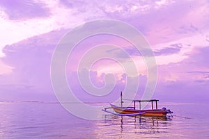 Traditional Balinese wooden fishing boat at dawn on Sanur beach in Bali. Cloudy pink dawn sky. Reflection in the water.