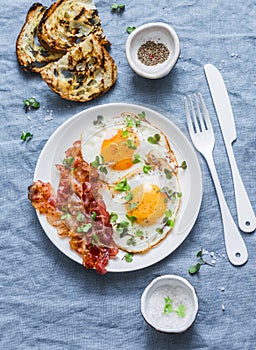 Traditional breakfast or snack - fried eggs, bacon, grilled bread on blue background, top view.