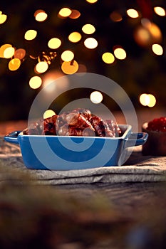 Traditional Dish Of Christmas Pigs In Blankets On Table Set For Meal With Tree Lights In Background