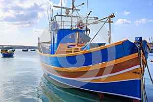 Traditional fishing boat, luzzu, anchored at Marsaxlokk, Malta. Blue sky with clouds and village background. Close up view.