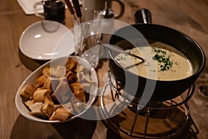 Traditional French cheese fondue in hot pot and white bowl with bread crackers. Utensil atop wooden table. Winter hot food.