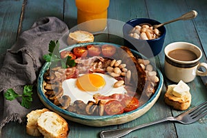 A traditional full English breakfast with fried egg, sausage, mushrooms, beans, bacon and tomatoes on a rustic wooden green table.