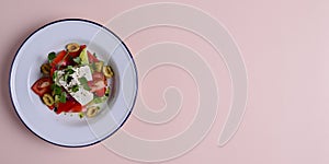 Traditional Greek salad with feta cheese and vegetables served in white plate over bright pastel pink background.