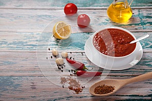 Traditional harissa in a porcelain cup, red hot chili pepper, lemon, oil, garlic and spices in a wooden spoon on a white