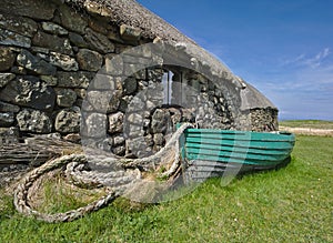 Traditional Hebridean blackhouse with thatched roof, wooden fishing boat and coiled rope.