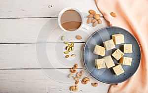 Traditional indian candy soan papdi in a blue ceramic plate with almond, pistache and a cup of coffee on a white wooden background