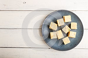 Traditional indian candy soan papdi in a blue ceramic plate with almond and pistache on a white wooden background. top view