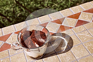 Traditional Italian dessert Tiramisu in a glass cup on a tile table in the garden. National cuisine recipe.