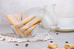 Traditional italian savoiardi or ladyfingers biscuits in a glass bowl and pieces of cookies and sugar on the table, a white cup of