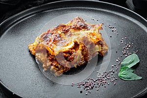 Traditional lasagna made with minced beef bolognese sauce topped with basil leafs, on plate, on black stone background