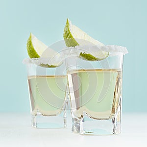 Traditional mexican golden tequila alcohol drink with lime and salt edge in shot glass on elegant blue color background, square.